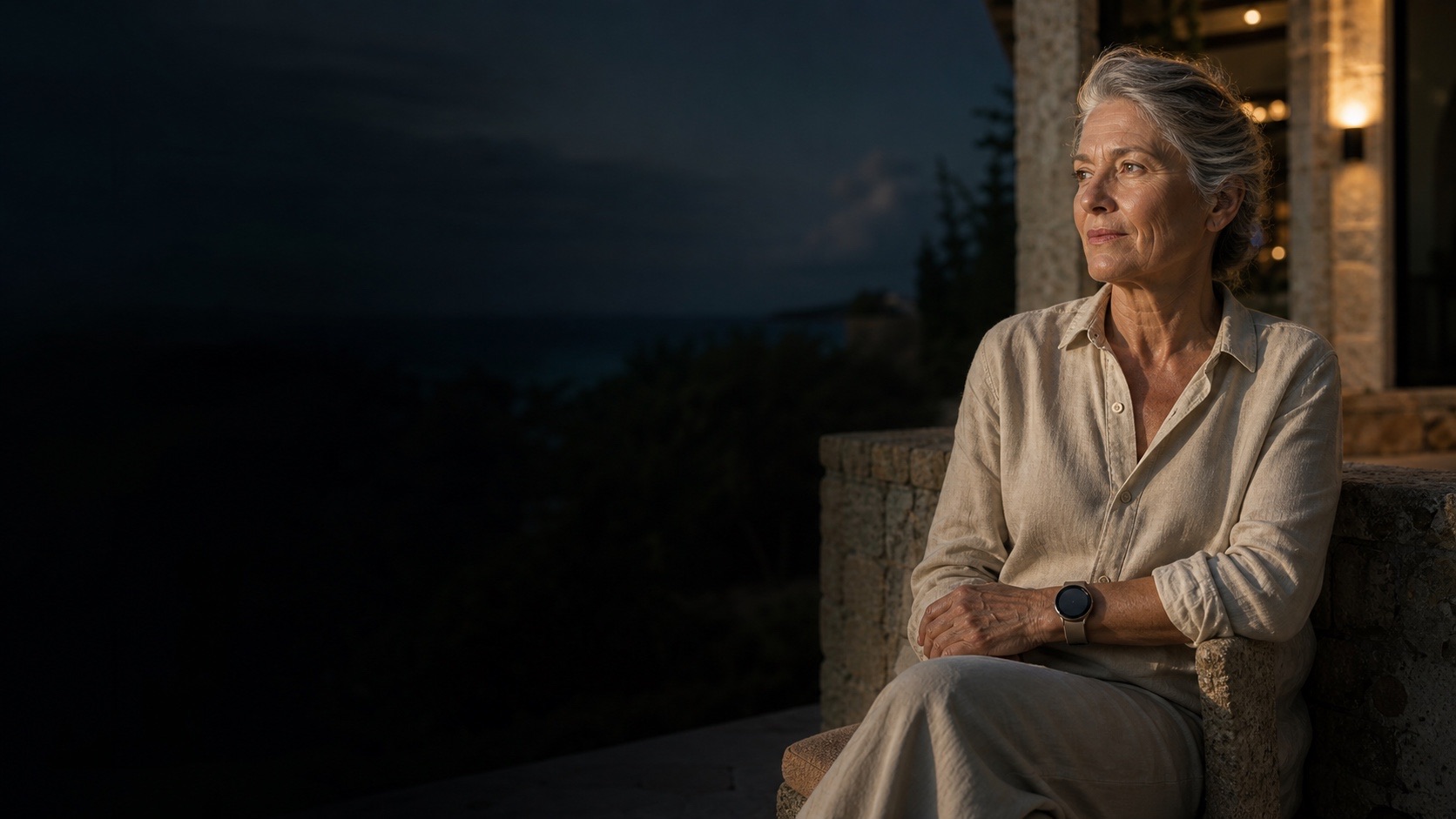 Eine selbstständige ältere Frau sitzt ruhig am Abend auf einer mediterranen Terrasse und trägt eine schlichte Smartwatch.